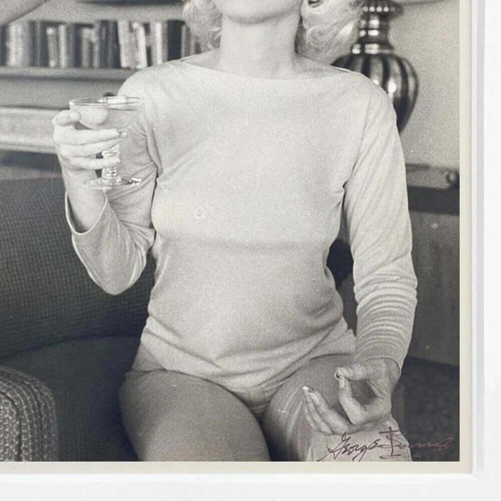 Vintage black and white photo of woman in long-sleeve top holding glass, seated on sofa