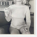 Vintage black and white photo of woman in long-sleeve top holding glass, seated on sofa