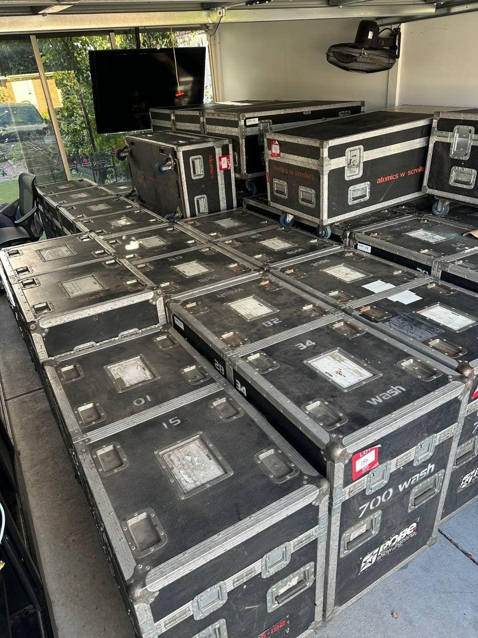 Stacked black road cases for stage lights, labeled '700 wash' and 'atomics w scrollers', in storage