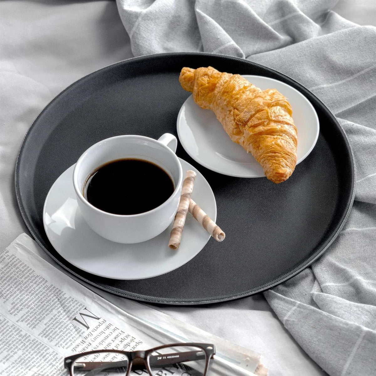 Black tray with coffee, croissant, and wafer sticks on bed with newspaper and glasses