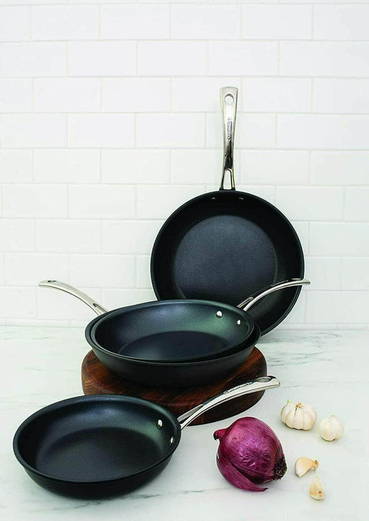 Set of three non-stick black frying pans with stainless steel handles on kitchen counter, onion and garlic beside them