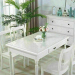 Elegant white dining table with clear PVC cover, surrounded by chairs and decorative plants, showcasing a modern dining space.