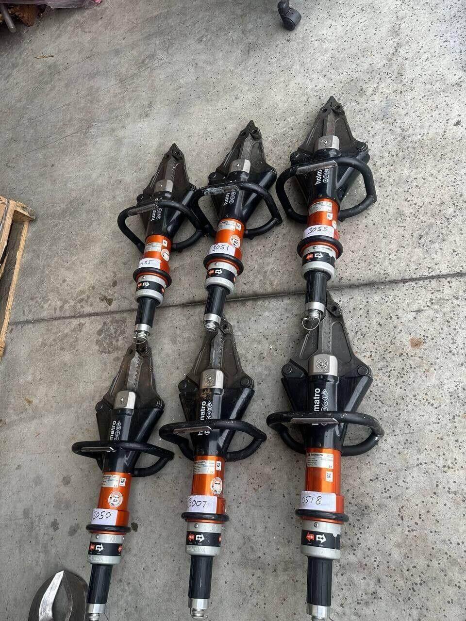 Six Holmatro rescue tools with orange and black design, labeled for inventory on concrete floor.