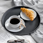 Black tray with coffee, croissant, and wafer sticks on bed with newspaper and glasses