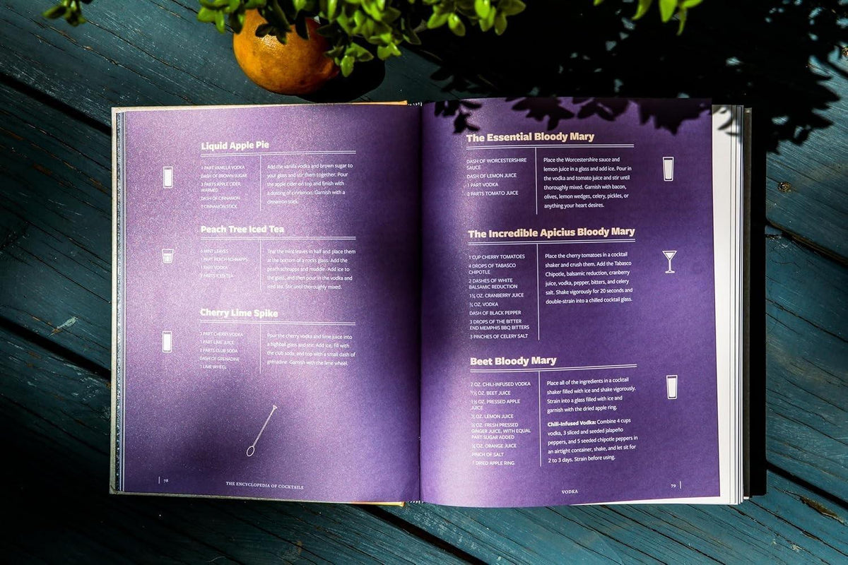 Cocktail recipe book open to Bloody Mary and vodka drink recipes on purple pages, styled on wooden table with orange and greenery