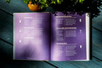 Cocktail recipe book open to Bloody Mary and vodka drink recipes on purple pages, styled on wooden table with orange and greenery