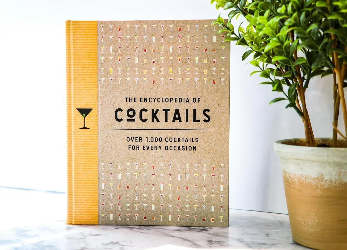 The Encyclopedia of Cocktails book with 1,000 recipes, displayed beside a potted plant on marble surface