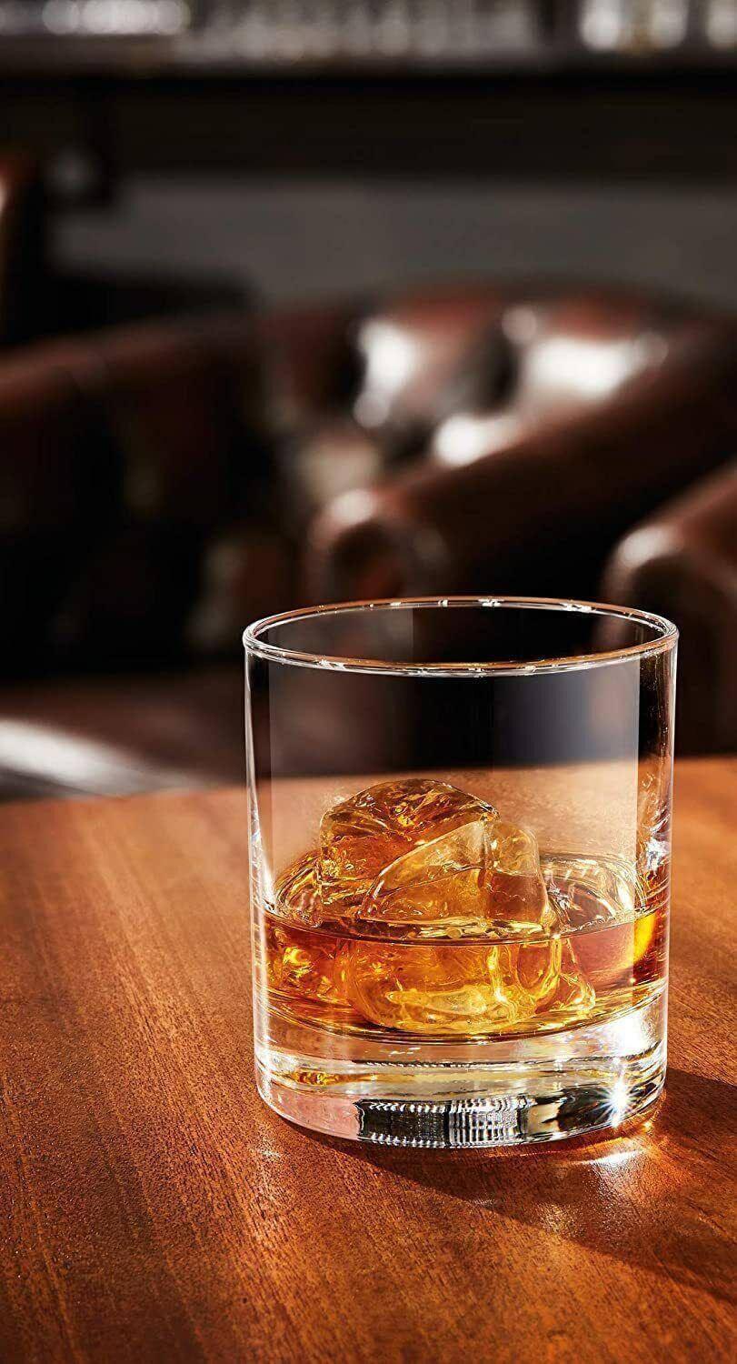 Clear tumbler whiskey glass with ice on wooden table, bar setting, drinkware