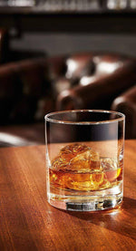 Clear tumbler whiskey glass with ice on wooden table, bar setting, drinkware