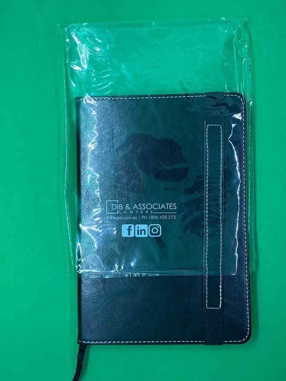 A5 black leather cover journal notebook with white stitching, partially in plastic, on green background.