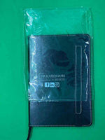 A5 black leather cover journal notebook with white stitching, partially in plastic, on green background.